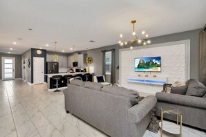 A modern living room with a grey sofa, a TV mounted on the wall, and a kitchen area in the background.