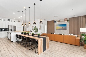 Une cuisine moderne avec un îlot central, des chaises noires, des luminaires suspendus et un grand téléviseur fixé au mur.