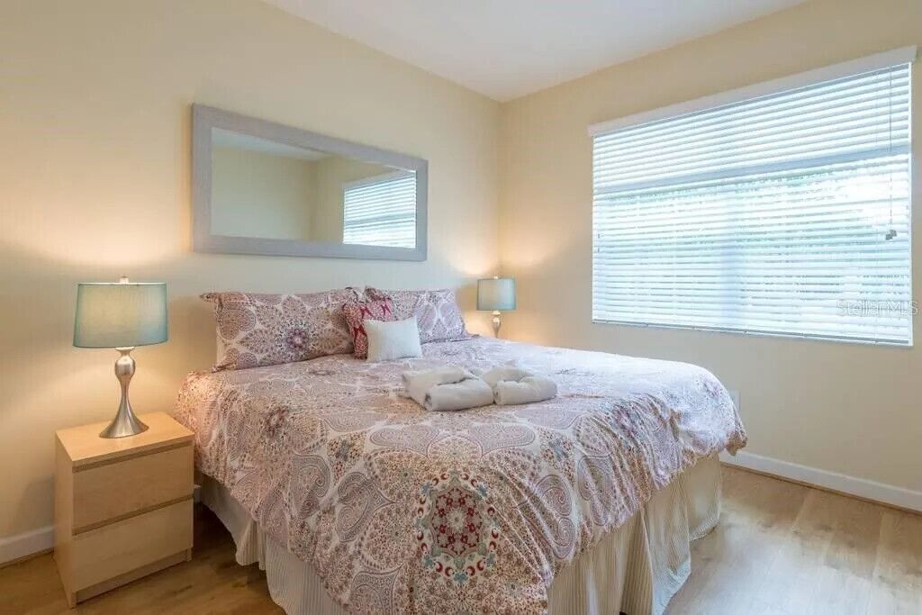 A bedroom with a large bed, two bedside lamps, a mirror above the bed, and a window with blinds.