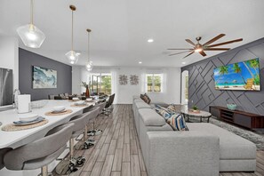 A modern living room with a grey sofa, a dining area with a long table, pendant lights, and a ceiling fan.