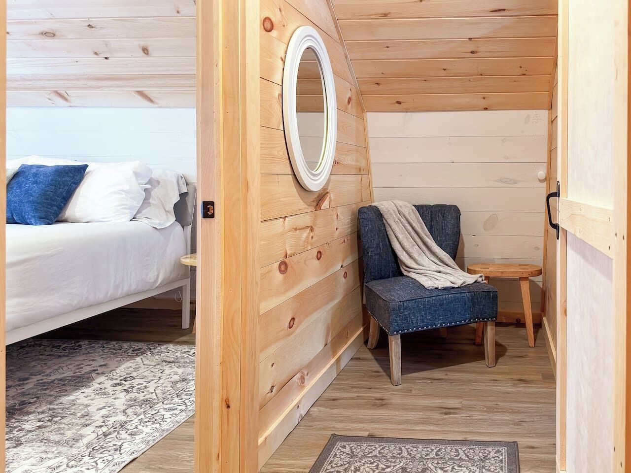 A cozy cabin interior with a bed, a chair, a small table, and a mirror on the wall.