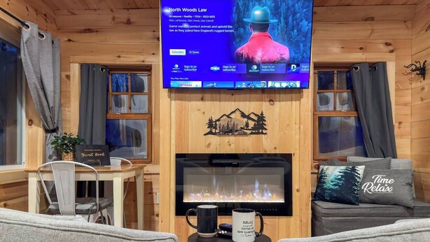 Een gezellig interieur van een blokhut met een televisie waarop 'North Woods Law' staat, een open haard, grijze banken en een kleine tafel met een mok.