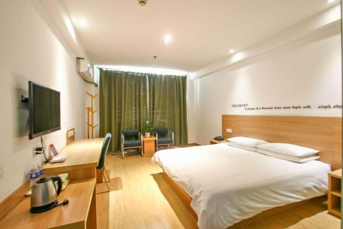 A hotel room with a bed, a desk with a kettle, a chair, a TV, and a window with curtains.