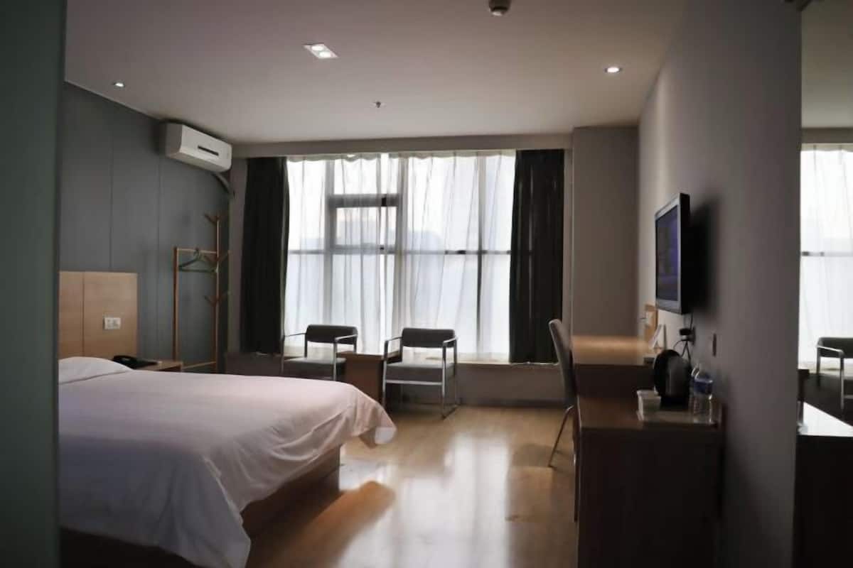 A hotel room with a bed, two chairs, a desk, a TV, and a large window with curtains.