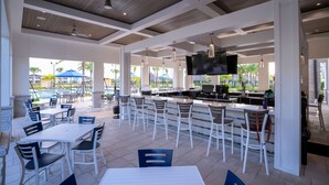 Un espace bar extérieur avec des tables et des chaises blanches, un téléviseur fixé au mur et une piscine visible au fond.