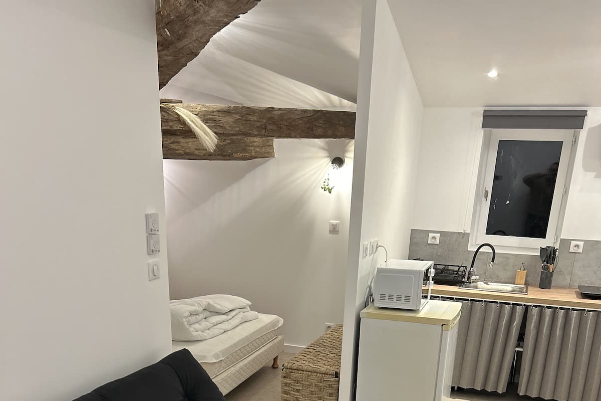Un espace de vie moderne comprenant un canapé noir, une petite table, un coin cuisine et un lit avec une couette blanche.