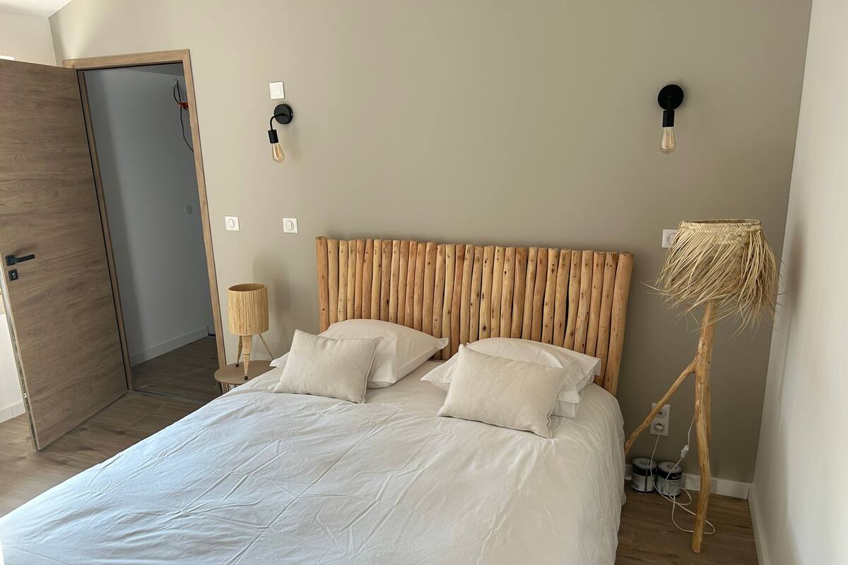 Une chambre à coucher avec une tête de lit en bois, un grand lit avec une literie blanche, deux appliques murales et une lampe de sol en osier.