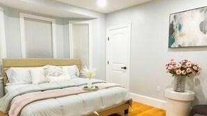 A bedroom with a bed, bedside tables, a nightstand, a window with blinds, a door, and a wall-mounted artwork. - 85a Charming, Cozy 3BR, 2BA Home Near Downtown Boston (Boston)
