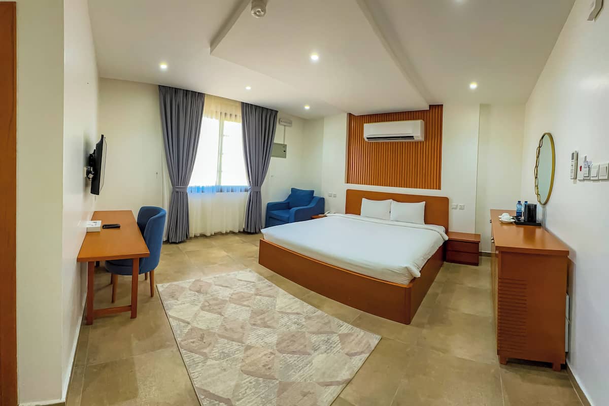 A modern hotel room with a large bed, a desk with a chair, a blue armchair, and a wooden dresser.