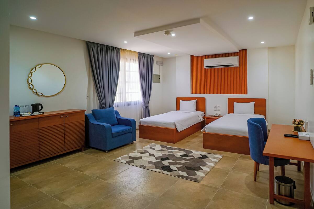 A hotel room with two beds, a desk, a chair, a coffee station, and a patterned rug.