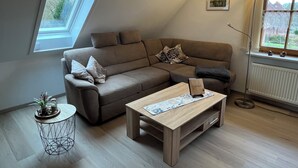 Een moderne woonkamer met een hoekbank, een salontafel, een bijzettafeltje met een plant en een groot raam dat voor natuurlijk licht zorgt.