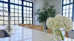 Cocina moderna con encimera de mármol blanco, pileta integrada y ventana grande con vista a árboles. Hay una planta en maceta y un florero con flores blancas sobre la encimera.
