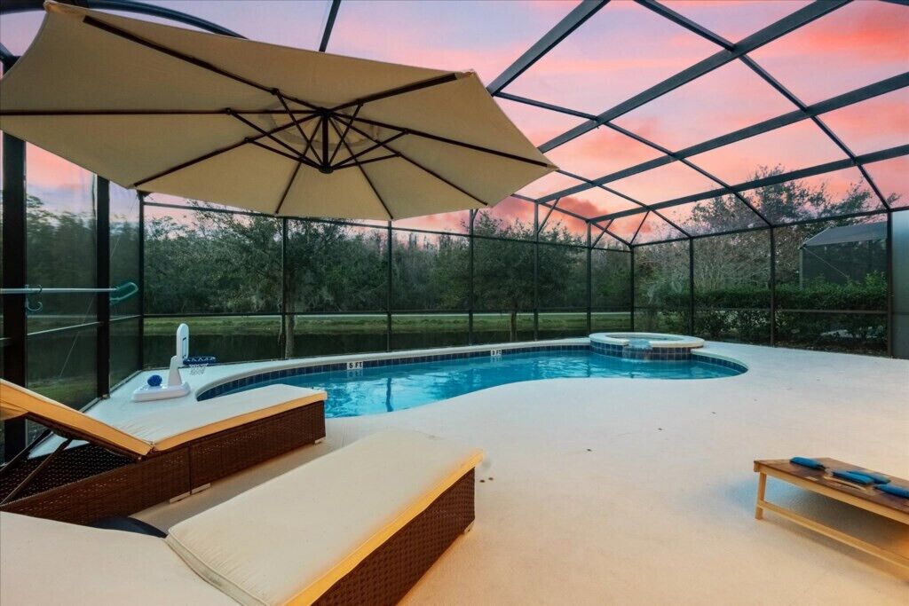 An indoor swimming pool with lounge chairs and a large umbrella, surrounded by glass walls and a metal frame structure.