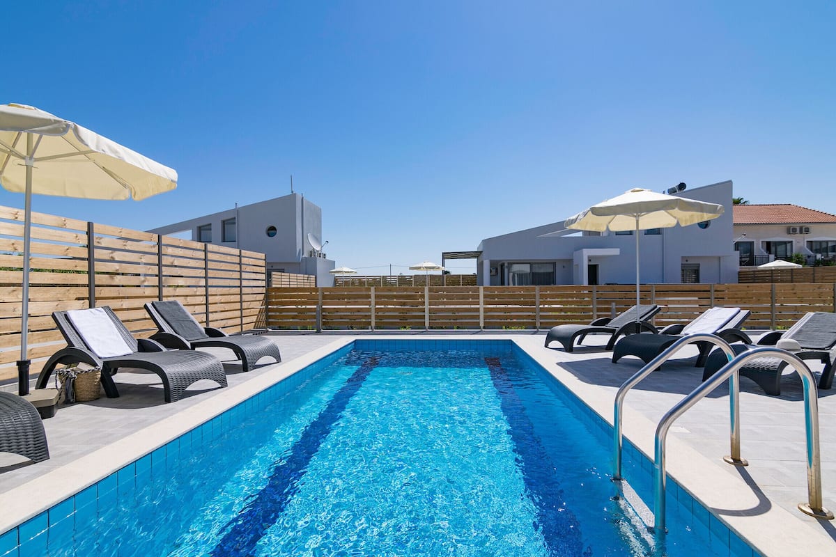 A swimming pool with lounge chairs and umbrellas, surrounded by a wooden fence and modern buildings.