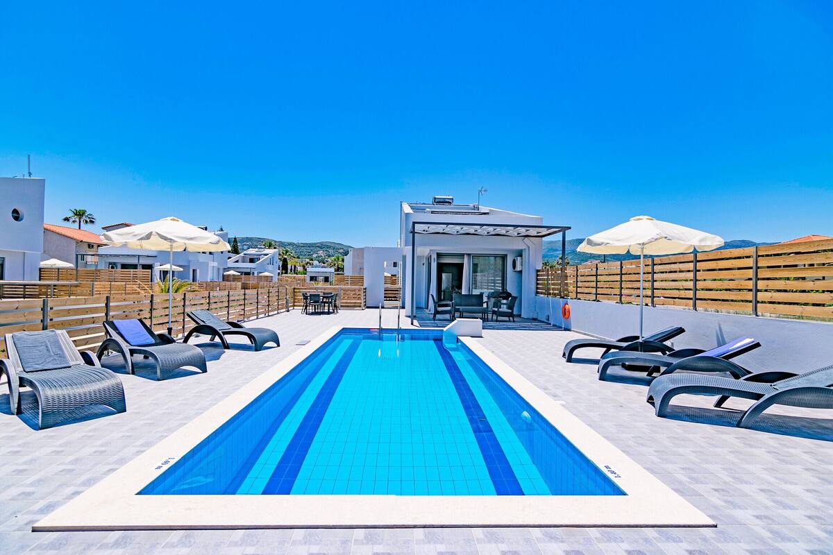 A rooftop swimming pool with lounge chairs, umbrellas, and a small building with a glass door.
