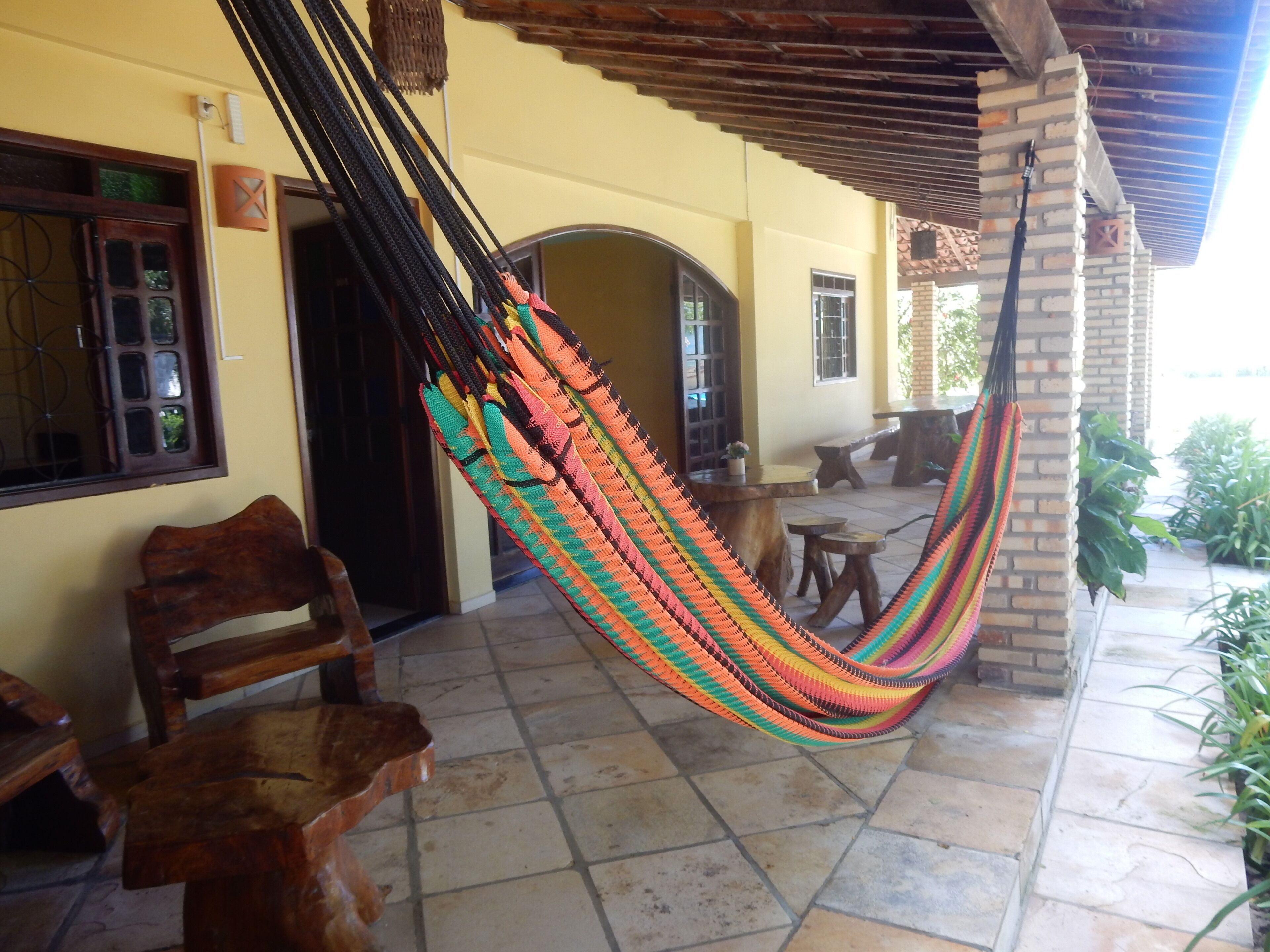 A hammock hanging between two brick pillars in a covered outdoor area with tiled flooring and wooden furniture.