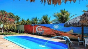A swimming pool with a mural, palm trees, and a thatched roof structure. - Pousada Iguana (Aracati)