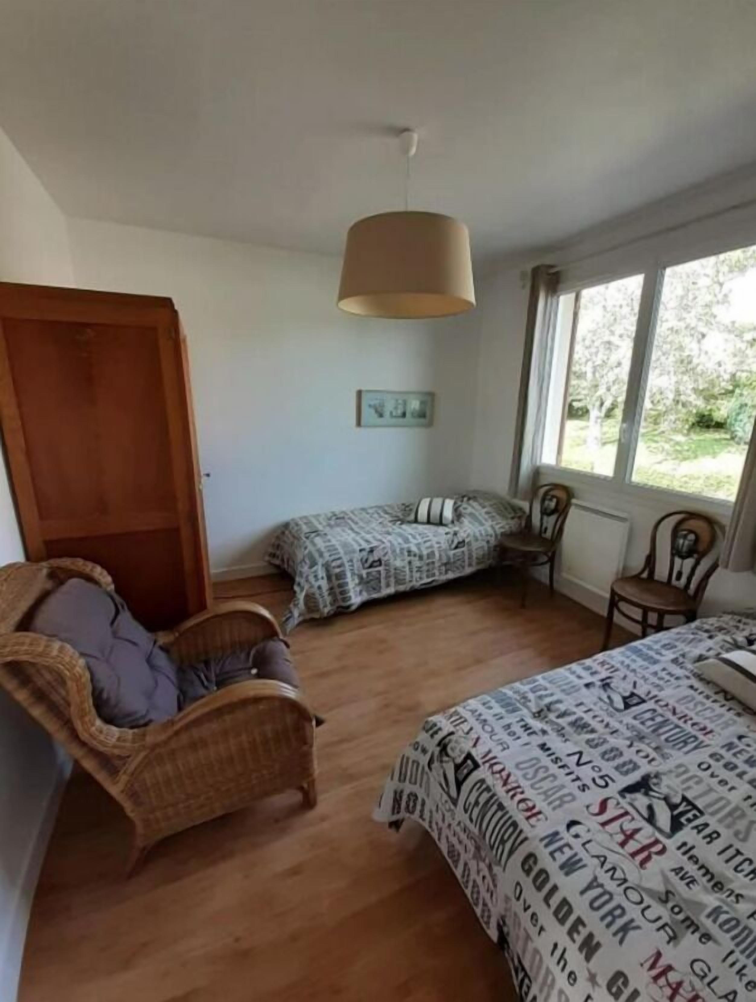 Un dormitorio con una cama, dos sillas, un sillón de mimbre, una lámpara colgante, una ventana y una puerta.