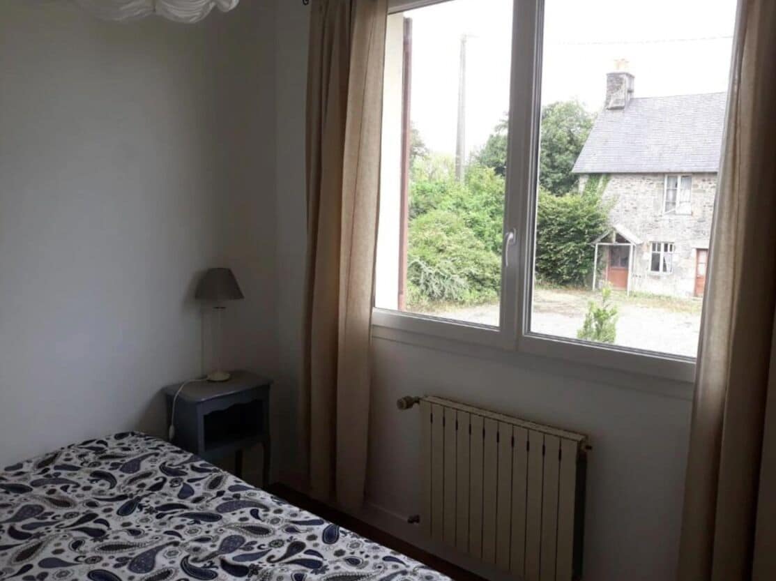 A bedroom with a bed, a bedside table with a lamp, a window with curtains, and a view of a house and greenery outside.