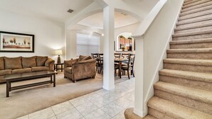 A spacious living room with a sofa, coffee table, and a dining area with chairs and a table. A staircase with carpeted steps is visible. - Last Minute Disney Getaway! Private Pool, Hot Tub & Game Room. Book Now! #65763 (Orlando)