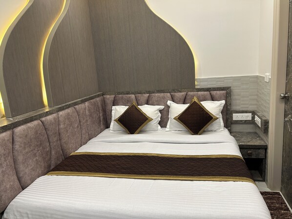 A modern hotel room with a large bed, a headboard with built-in lighting, a bedside table, and a power outlet. - Hotel Shree Guru (New Delhi)