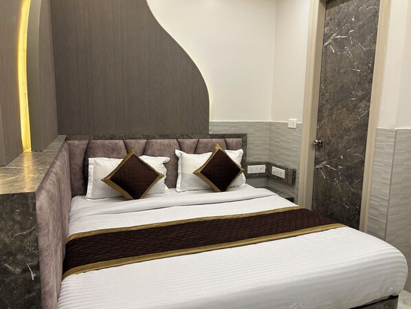 A modern bedroom with a curved headboard, a marble accent wall, and a bed with white bedding and brown pillows. - Hotel Shree Guru (New Delhi)