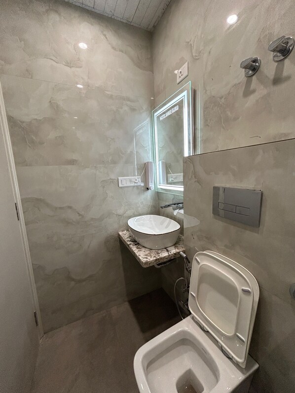 A modern bathroom with a marble wall, a white sink, a mirror, and a toilet. - Hotel Shree Guru (New Delhi)