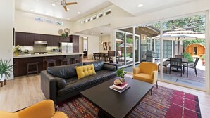 Un salon moderne comprenant un canapé, une table basse et des fauteuils, un coin cuisine avec des meubles sombres, et une grande porte-fenêtre coulissante donnant sur une terrasse extérieure.