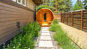 Un sauna en forme de tonneau en bois, accessible par un chemin de pierres, entouré de verdure et délimité par une clôture en bois.