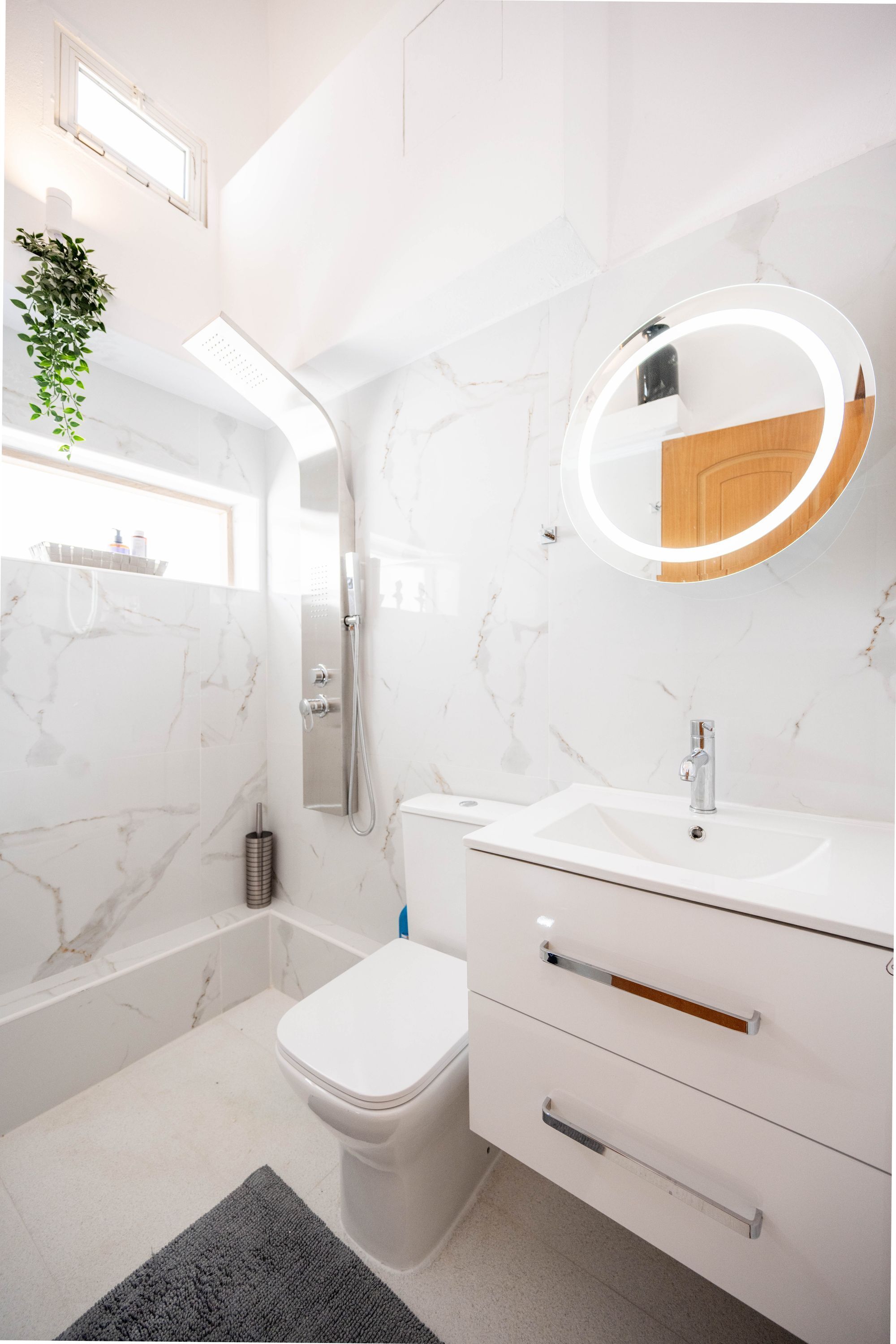 A modern bathroom with a white marble wall, a round mirror with a light, a white sink, and a toilet.
