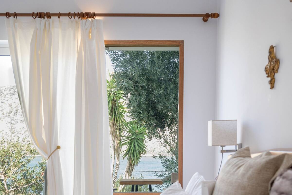 A bedroom with a bed, white curtains, a window with a view of palm trees, and a bedside table with a lamp.