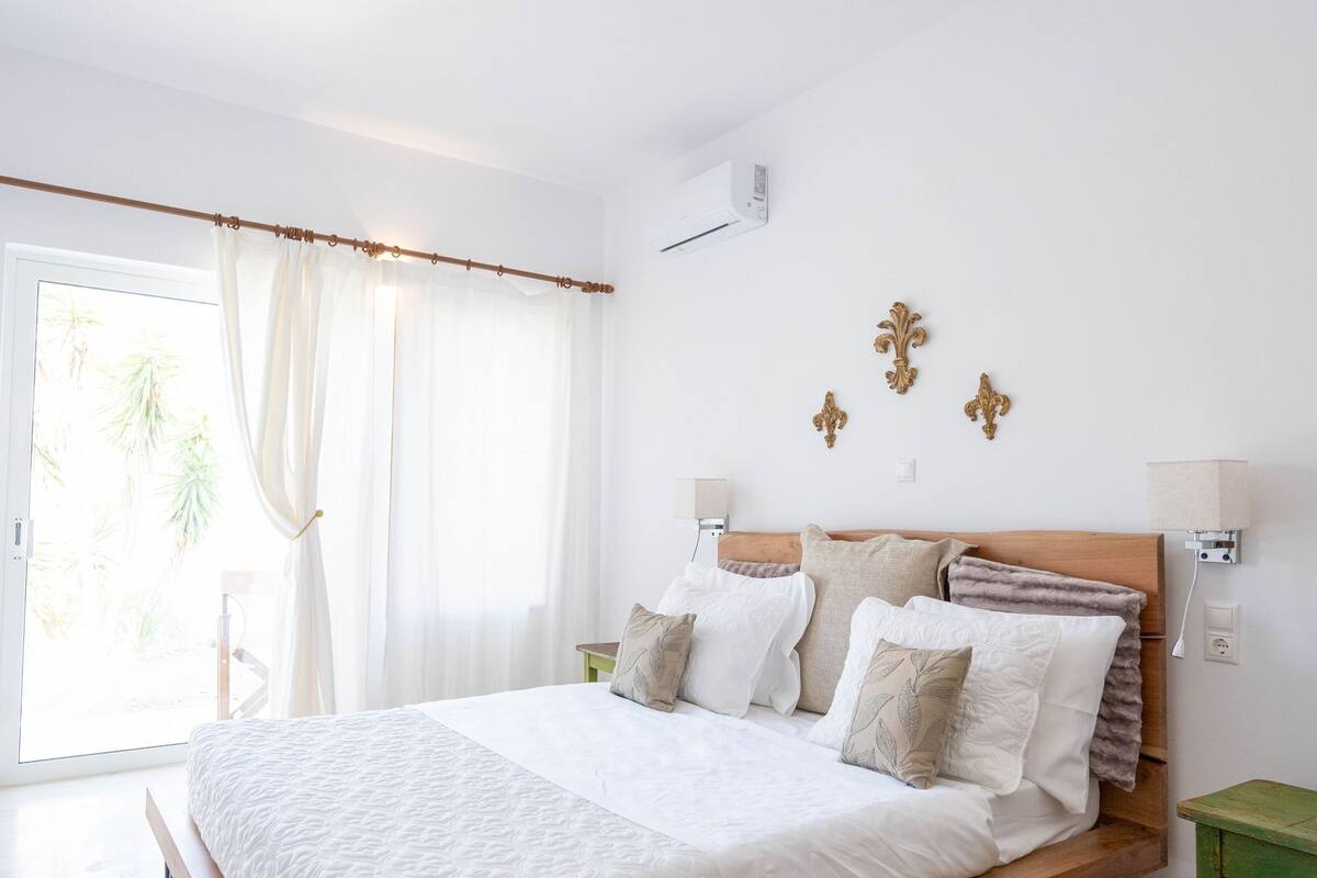 A bedroom with a wooden bed, white bedding, a green nightstand, and a view of palm trees through the window.