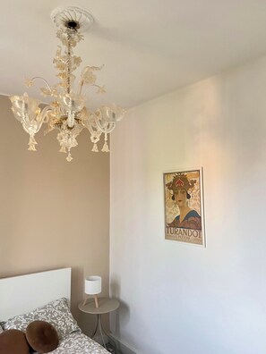 Ein Schlafzimmer mit einem Kronleuchter, einem gerahmten Poster, einem Nachttisch mit Lampe und gemusterten Betttextilien.