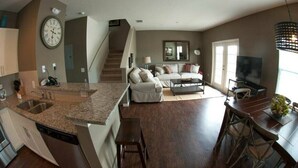 A modern kitchen with granite countertops, a dishwasher, and a sink. There is a dining area with a wooden table and chairs. A living room with a sofa and a television is visible in the background. - Last Minute Disney Getaway! Townhouse with Private Pool. Book Now! #125775 (Orlando)