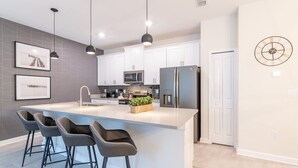 A modern kitchen with a central island, bar stools, a microwave, a refrigerator, and a clock on the wall.