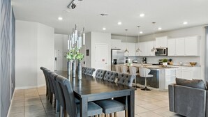 A modern kitchen with white cabinets, a central island, and a dining area with a dark wood table and grey upholstered chairs. - Last Minute Disney! Private Pool, Hot Tub, Game Room & Themed Bedrooms. Book Now! #128641 (Orlando)