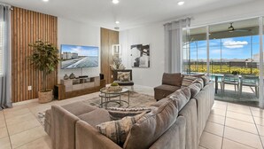 A modern living room with a large flat-screen TV, a sectional sofa, a coffee table, and a view of a pool and mountains through large windows. - Last Minute Disney! Private Pool, Hot Tub, Game Room & Themed Bedrooms. Book Now! #128641 (Orlando)