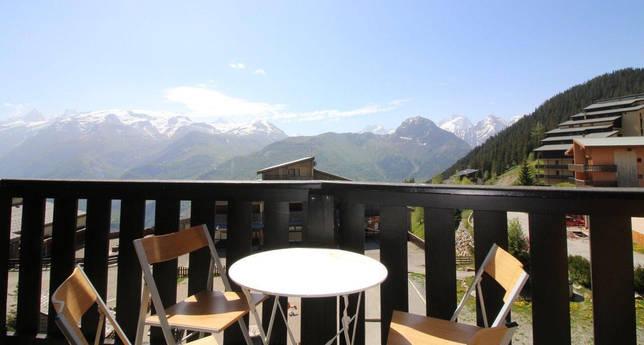 A balcony with a table and chairs, overlooking a mountainous landscape with snow-capped peaks and a forested area.