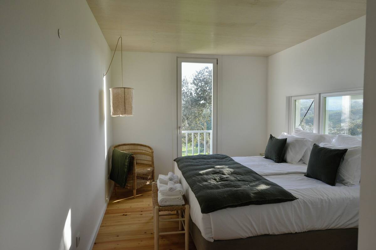 Quarto com uma cama grande, uma cadeira, uma mesinha com toalhas e uma janela com vista para árvores.