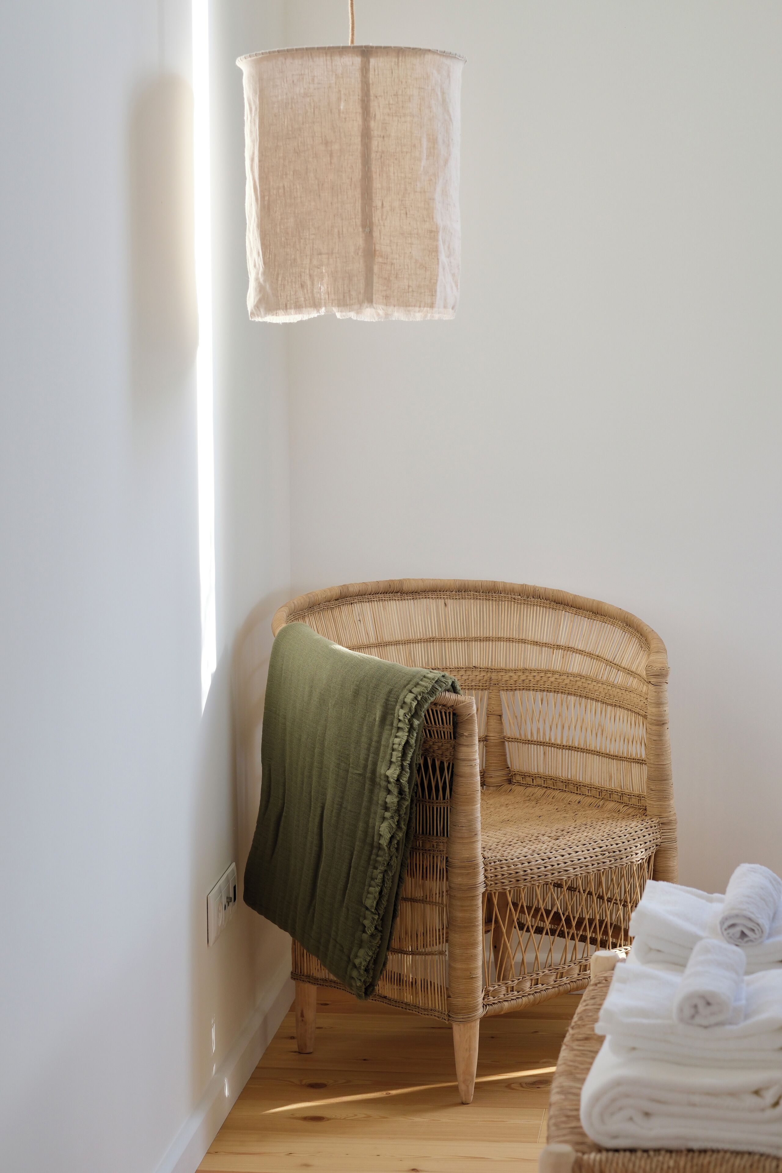 Una silla de mimbre con una manta verde, una pila de toallas blancas y una lámpara colgante en una habitación con paredes blancas y suelo de madera.