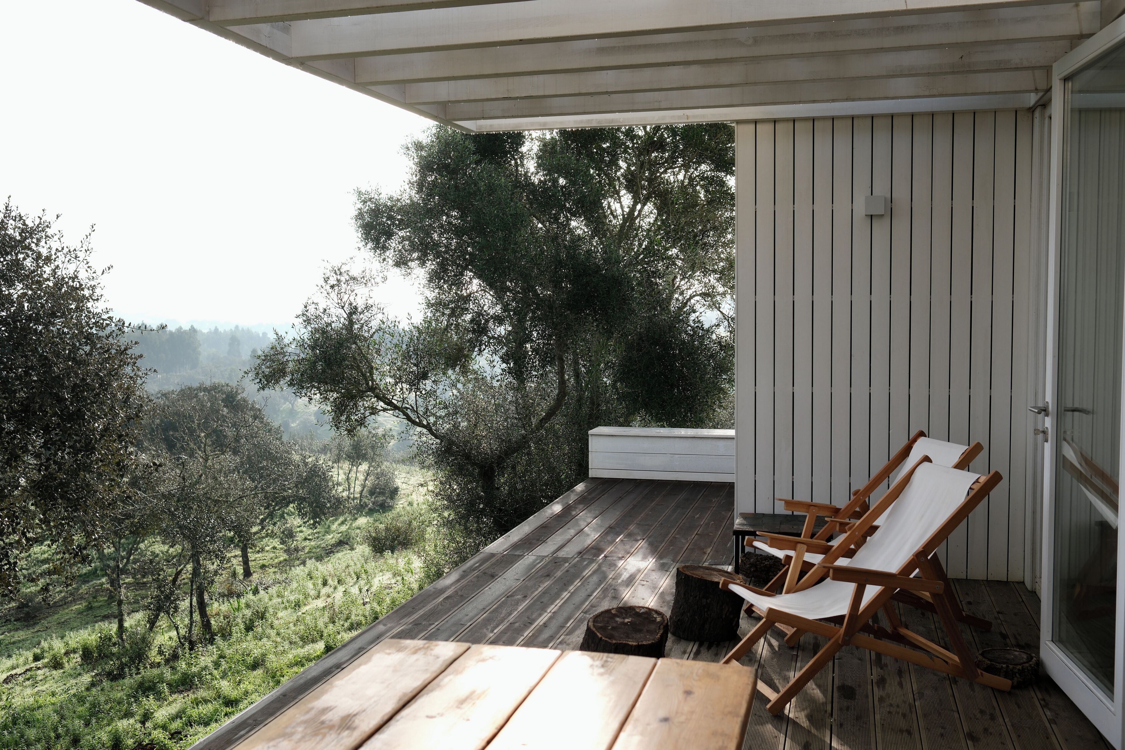A wooden deck with two white lounge chairs and a small table, overlooking a lush landscape with trees and hills.