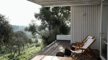 A wooden deck with two white lounge chairs and a small table, overlooking a lush landscape with trees and hills.
