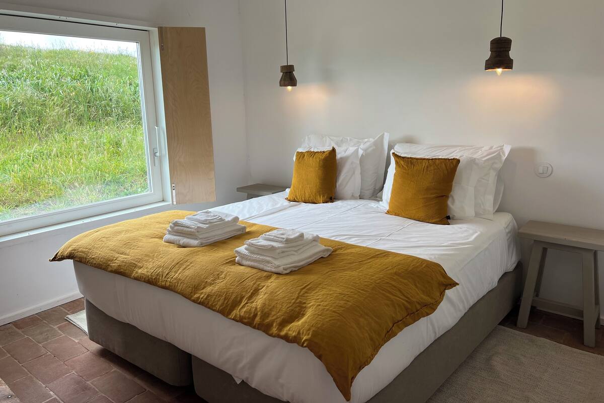 Quarto com cama grande, duas mesas de cabeceira, janela com vista para área verde e duas luminárias pendentes.