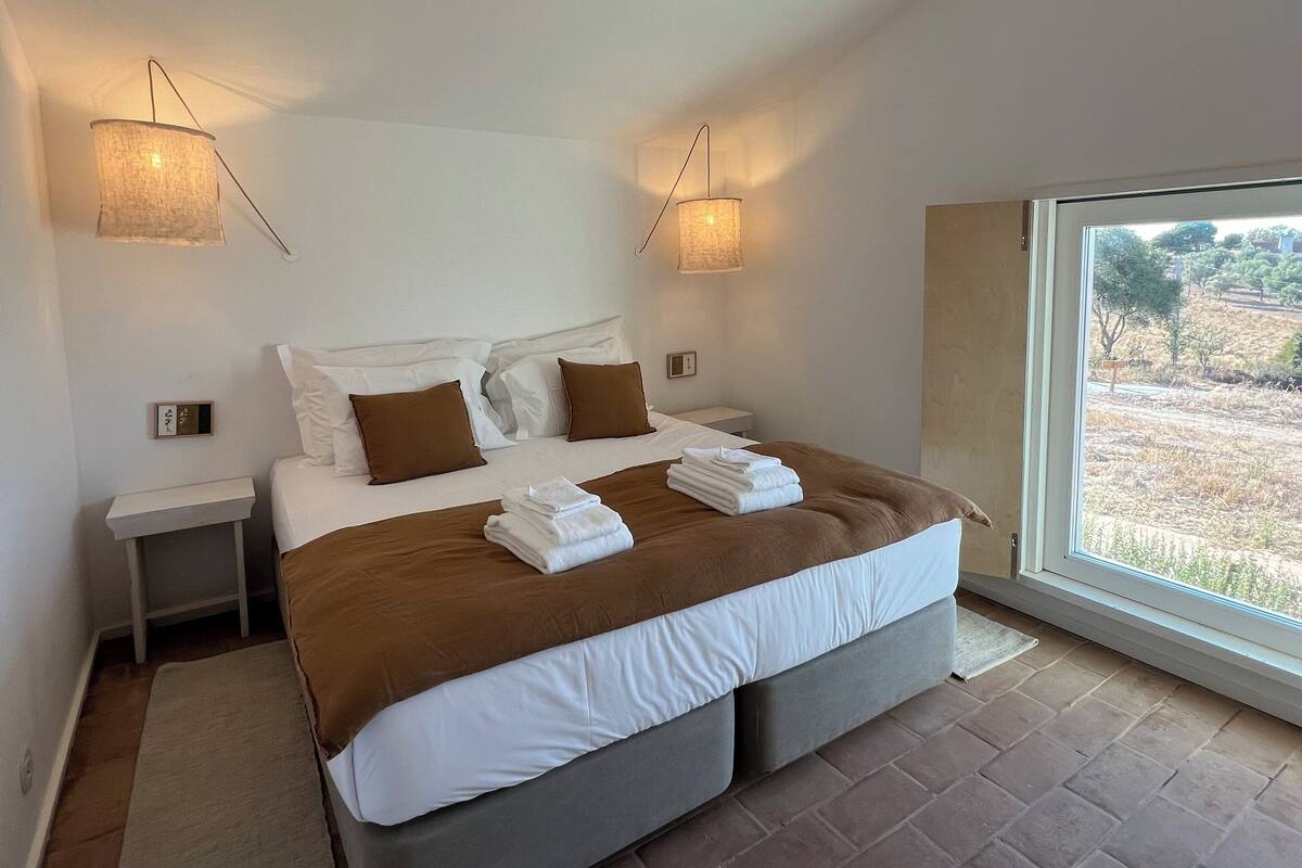 Quarto moderno com uma cama grande, duas mesas de cabeceira, um criado-mudo e uma janela com vista para o exterior.