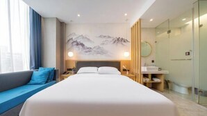 Une chambre d’hôtel comprenant un lit, un canapé, une table de chevet, une commode, un miroir et une salle de bain avec un lavabo et une douche en verre.
