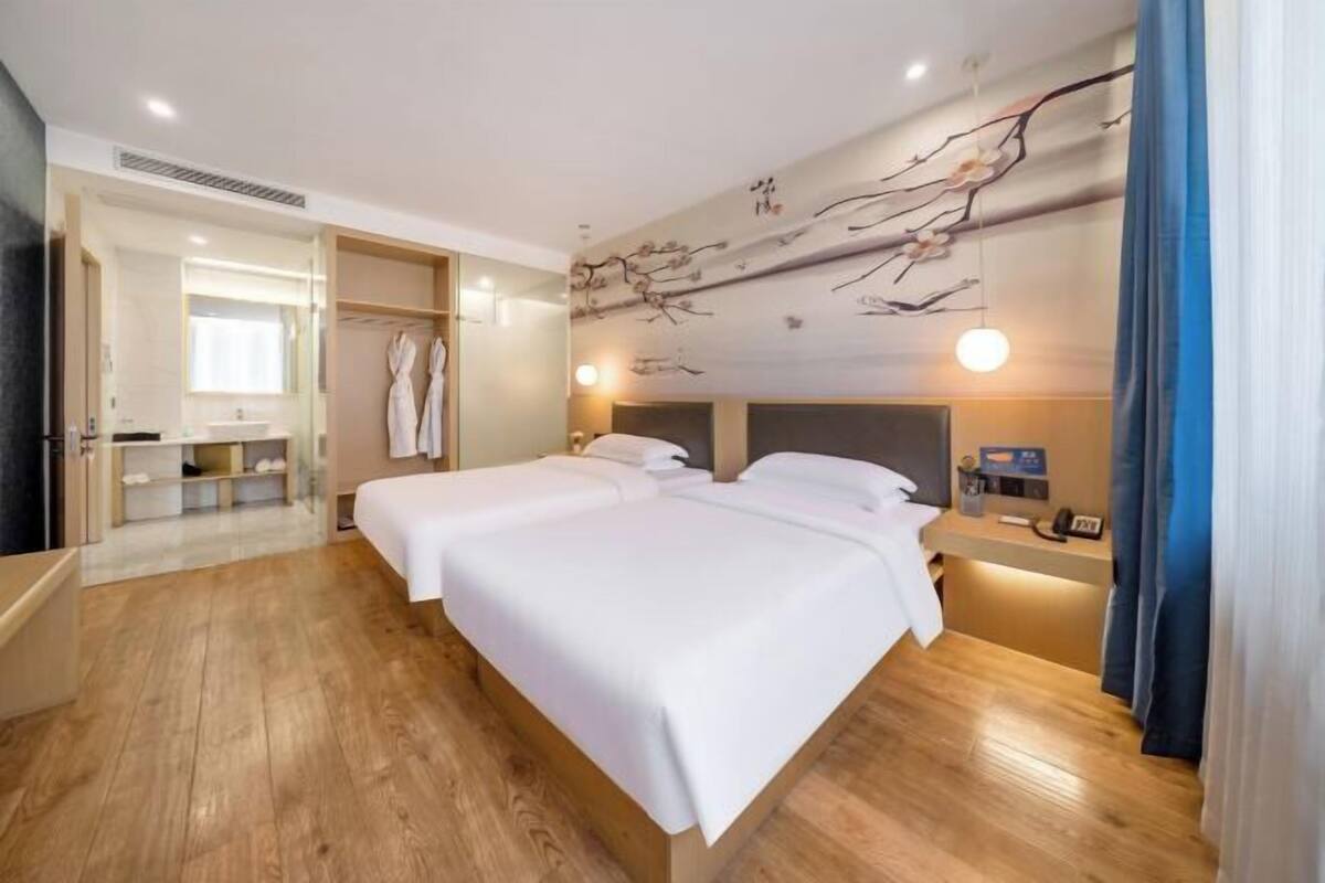 A hotel room with a wooden floor, two beds, a bedside table, a mirror, a robe rack, and a wall with a branch design.