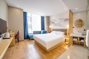 Et moderne hotelværelse med en stor seng, et skrivebord, en stol, et fjernsyn, et vindue med gardiner, et badeværelse med vask og spejl, og en væg med et bjergmaleri.