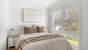 A bedroom with a bed, a nightstand, a lamp, a window with blinds, and a wall-mounted artwork. - Piccolo Cottage - Firepit - 4 Mins Walk to Beach (Callala Beach)