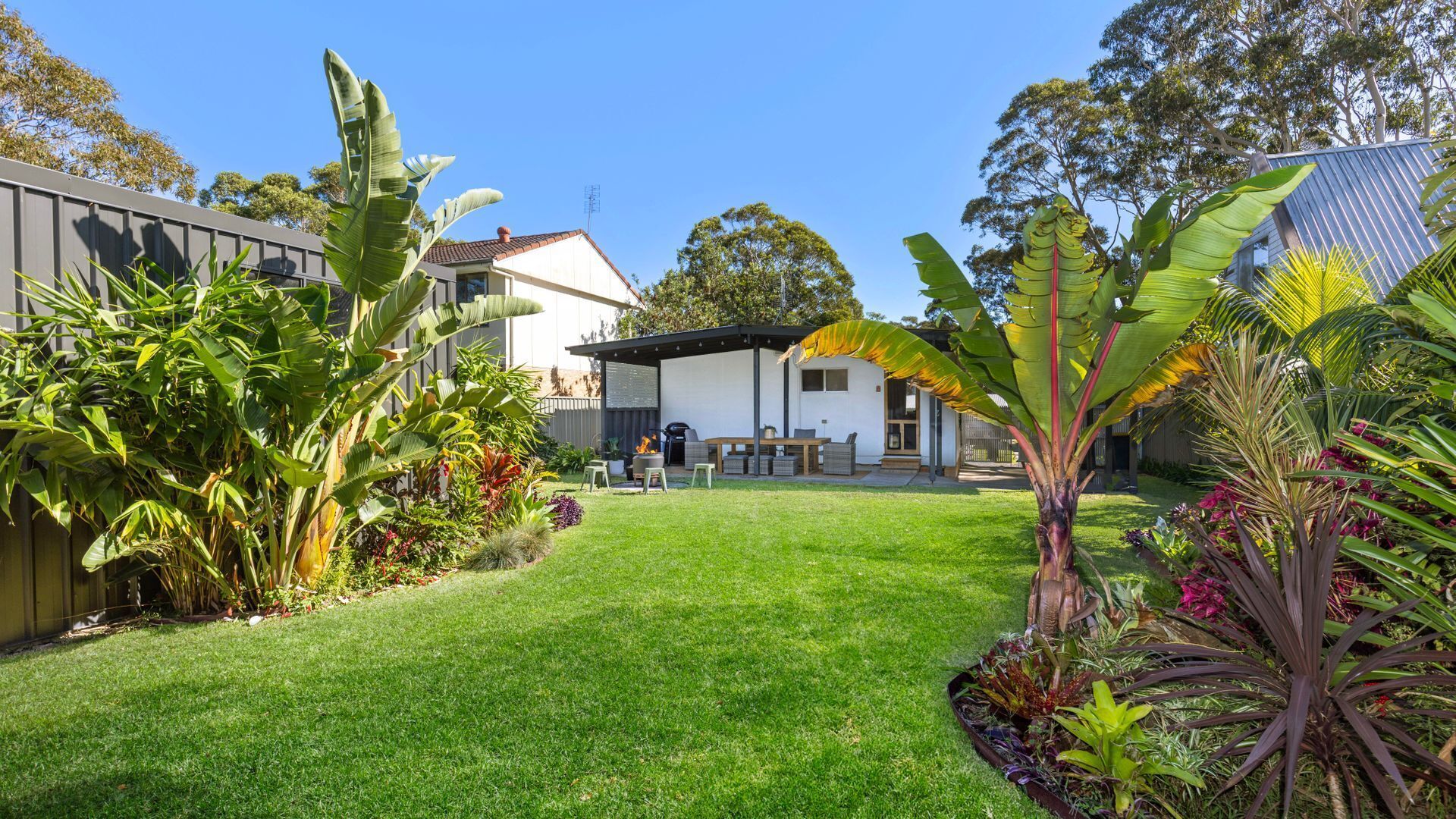 A well-maintained backyard with a grassy lawn, a barbecue area, and various tropical plants.