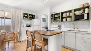 A modern kitchen with white cabinetry, a wooden island, wicker chairs, and green shelves with dishes. - Piccolo Cottage - Firepit - 4 Mins Walk to Beach (Callala Beach)